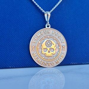 K&ouml;nnte beinhalten: Silberkette mit einem runden Anh&auml;nger, der einen Sch&auml;del mit Zahnr&auml;dern und die Inschrift "TEMPUS FUGIT MEMENTO MORI" in einem Kreis zeigt.