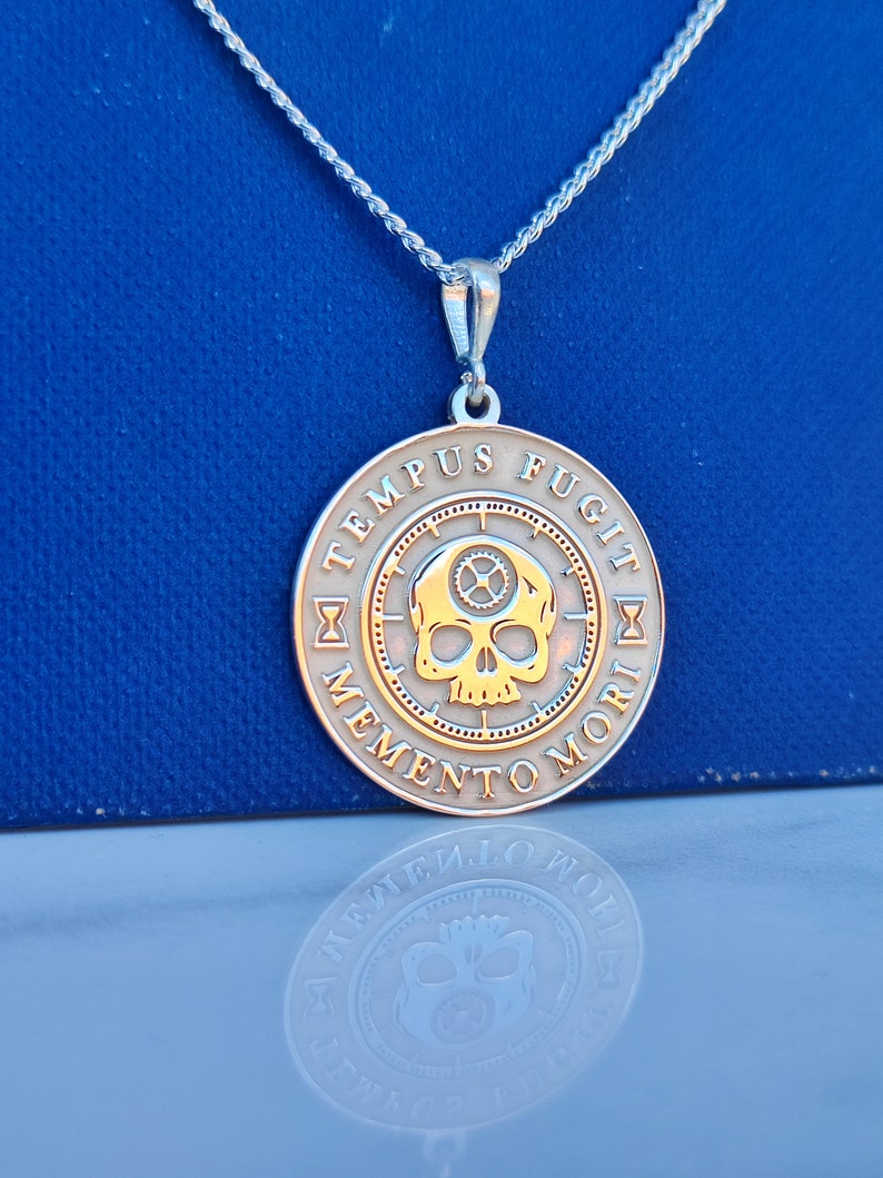 K&ouml;nnte beinhalten: Silberkette mit einem runden Anh&auml;nger, der einen Sch&auml;del mit Zahnr&auml;dern und die Inschrift "TEMPUS FUGIT MEMENTO MORI" in einem Kreis zeigt.