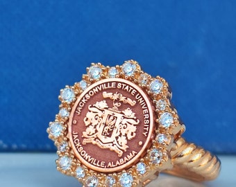 Könnte beinhalten: Ein goldener Ring mit einem braunen und goldenen Emblem mit den Worten "Jacksonville State University" und "Jacksonville, Alabama". Das Emblem ist von einem Kreis aus klaren Strasssteinen umgeben.
