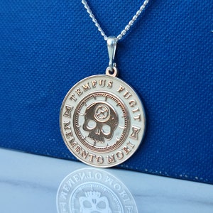 K&ouml;nnte beinhalten: Eine silberne Halskette mit einem runden Anh&auml;nger, der einen Totenkopf und die Inschrift "Memento Mori, Tempus Fugit" zeigt. Der Anh&auml;nger ist zweifarbig, mit einem silbernen Au&szlig;enring und einem goldenen Innenkreis.
