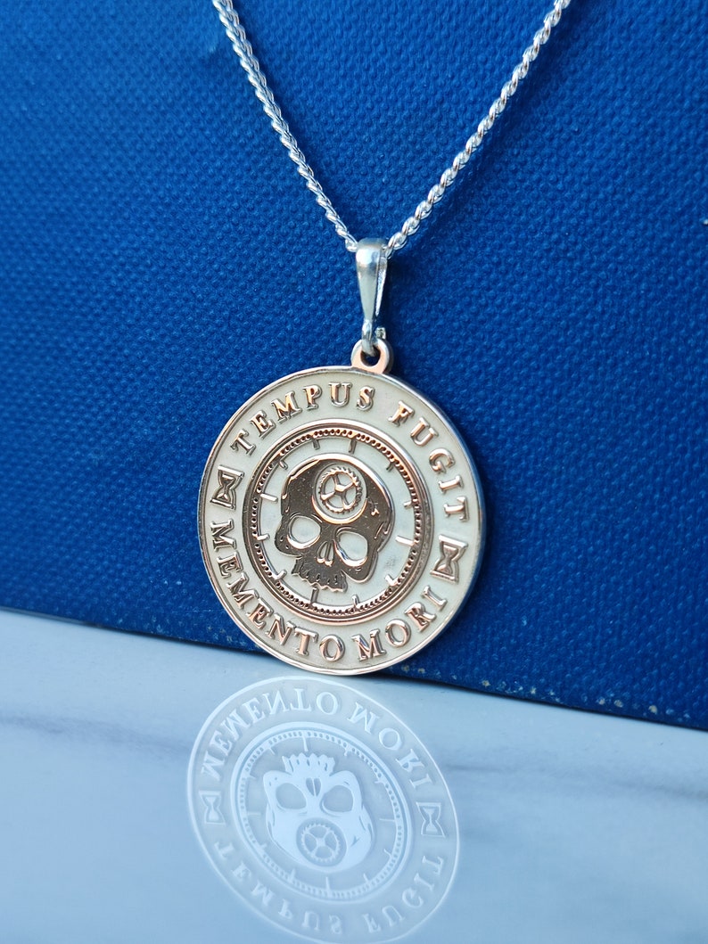 K&ouml;nnte beinhalten: Eine silberne Halskette mit einem runden Anh&auml;nger, der einen Totenkopf und die Inschrift "Memento Mori, Tempus Fugit" zeigt. Der Anh&auml;nger ist zweifarbig, mit einem silbernen Au&szlig;enring und einem goldenen Innenkreis.