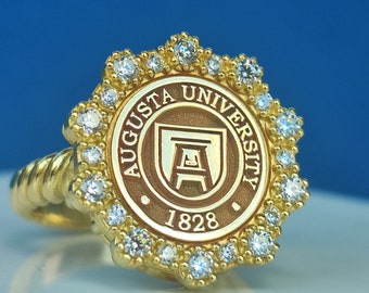 Könnte beinhalten: Ein goldener Ring mit einem braunen und goldenen Logo der Augusta University und einem Kreis aus kleinen Diamanten am Rand.