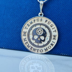 K&ouml;nnte beinhalten: Eine silberne Halskette mit einem runden Anh&auml;nger, der einen Sch&auml;del mit Zahnr&auml;dern und den Text "TEMPUS FUGIT MEMENTO MORI" in einem Kreis zeigt.