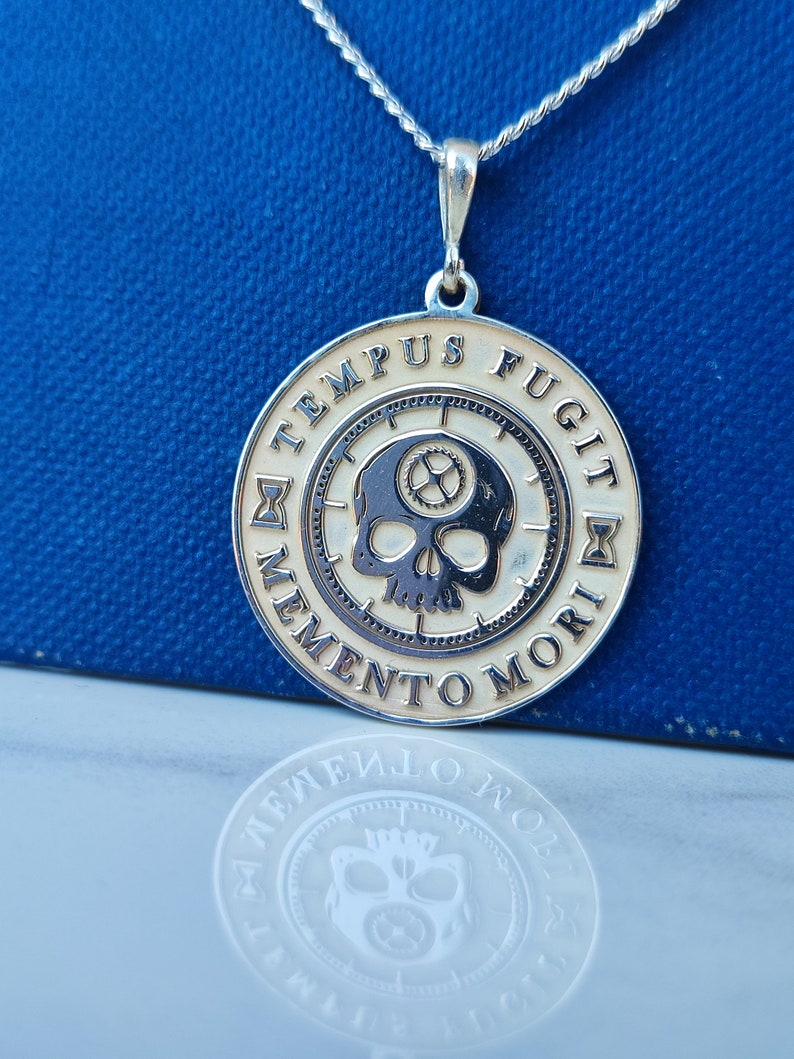 K&ouml;nnte beinhalten: Eine silberne Halskette mit einem runden Anh&auml;nger, der einen Sch&auml;del mit Zahnr&auml;dern und den Text "TEMPUS FUGIT MEMENTO MORI" in einem Kreis zeigt.