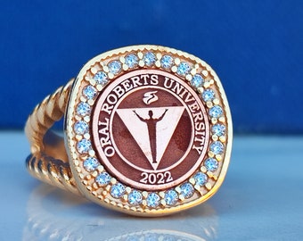 Könnte beinhalten: Ein goldener Ring mit einem roten und goldenen kreisförmigen Design mit den Worten "Oral Roberts University" und dem Jahr "2022". Der Ring ist mit einer Reihe von klaren Edelsteinen verziert.