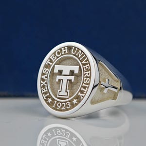 May include: A silver ring with a round, engraved top featuring the Texas Tech University logo and the year 1923. The ring has a cross design on the side.