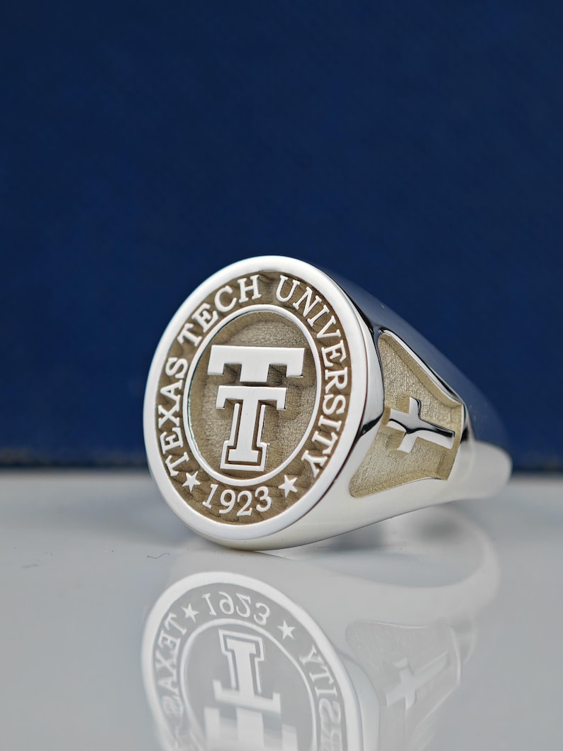 May include: A silver ring with a round, engraved top featuring the Texas Tech University logo and the year 1923. The ring has a cross design on the side.