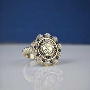 May include: A gold ring with a blue and white stone setting. The ring features the Auburn University seal with the text "Auburn University" and "For the Advancement of Science and Arts" around the seal. The date "05.11.2025" is inscribed below the seal.
