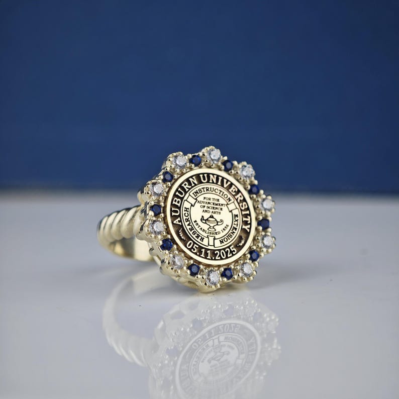 May include: A gold ring with a blue and white stone setting. The ring features the Auburn University seal with the text "Auburn University" and "For the Advancement of Science and Arts" around the seal. The date "05.11.2025" is inscribed below the seal.