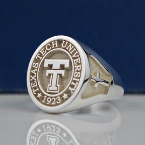 May include: A silver ring with a circular design featuring the words "Texas Tech University" and a stylized "T" with the year "1923" below. The ring has a cross design on the side.