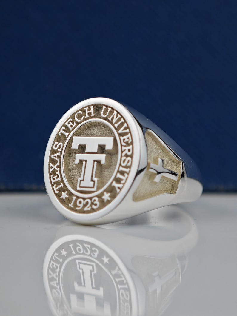 May include: A silver ring with a circular design featuring the words "Texas Tech University" and a stylized "T" with the year "1923" below. The ring has a cross design on the side.