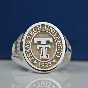 May include: A silver ring with a raised circular design featuring the words "Texas Tech University" and the year "1923" surrounding a stylized "T".