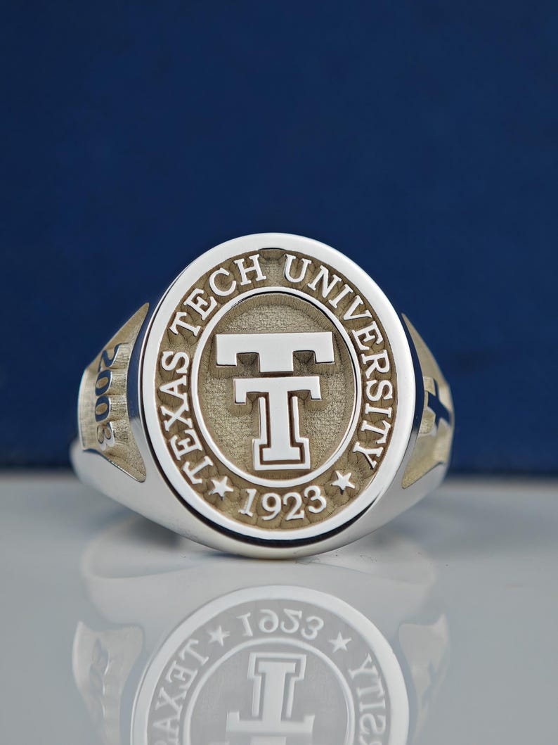 May include: A silver ring with a raised circular design featuring the words "Texas Tech University" and the year "1923" surrounding a stylized "T".