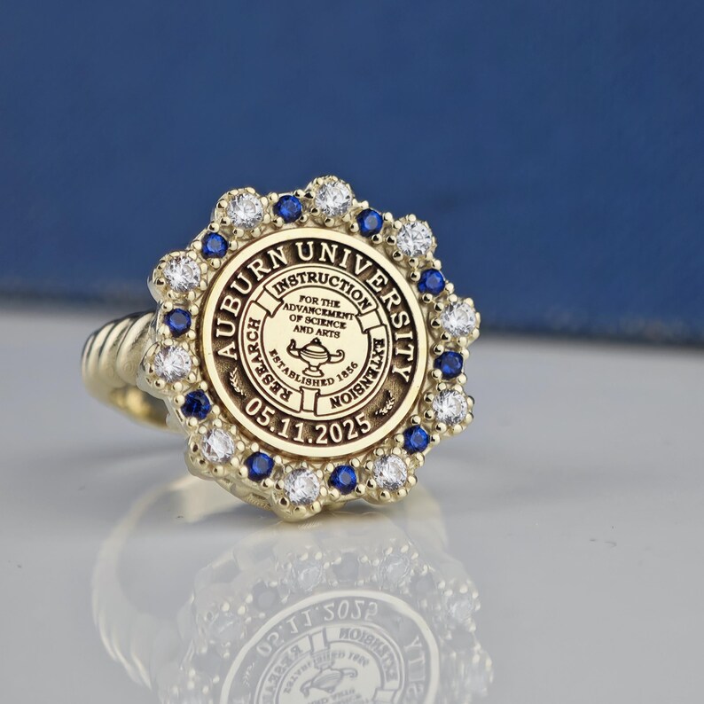 May include: A gold ring with a blue and white stone setting. The ring features the Auburn University seal with the text "Auburn University, Instruction, Research, Extension, For the Advancement of Science and Arts, Established 1856, 05.11.2025".