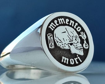 May include: A silver signet ring with a black oval top featuring a skull and crossbones design. The text "Memento Mori" is inscribed around the skull.