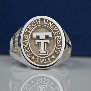 May include: A silver ring with a gold-toned center featuring the Texas Tech University logo and the year 1923.
