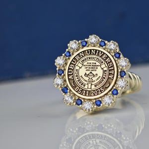 May include: A gold ring with a blue and white stone setting. The ring features the Auburn University seal with the text "Auburn University" and "Instruction, Research, Extension" around the edge. The seal also includes the text "For the Advancement of Science and Arts" and "Established 1856". The date "05.11.2025" is inscribed below the seal.