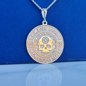 K&ouml;nnte beinhalten: Silberkette mit einem runden Anh&auml;nger, der einen Sch&auml;del mit Zahnr&auml;dern und die Inschrift "TEMPUS FUGIT MEMENTO MORI" in einem Kreis zeigt.