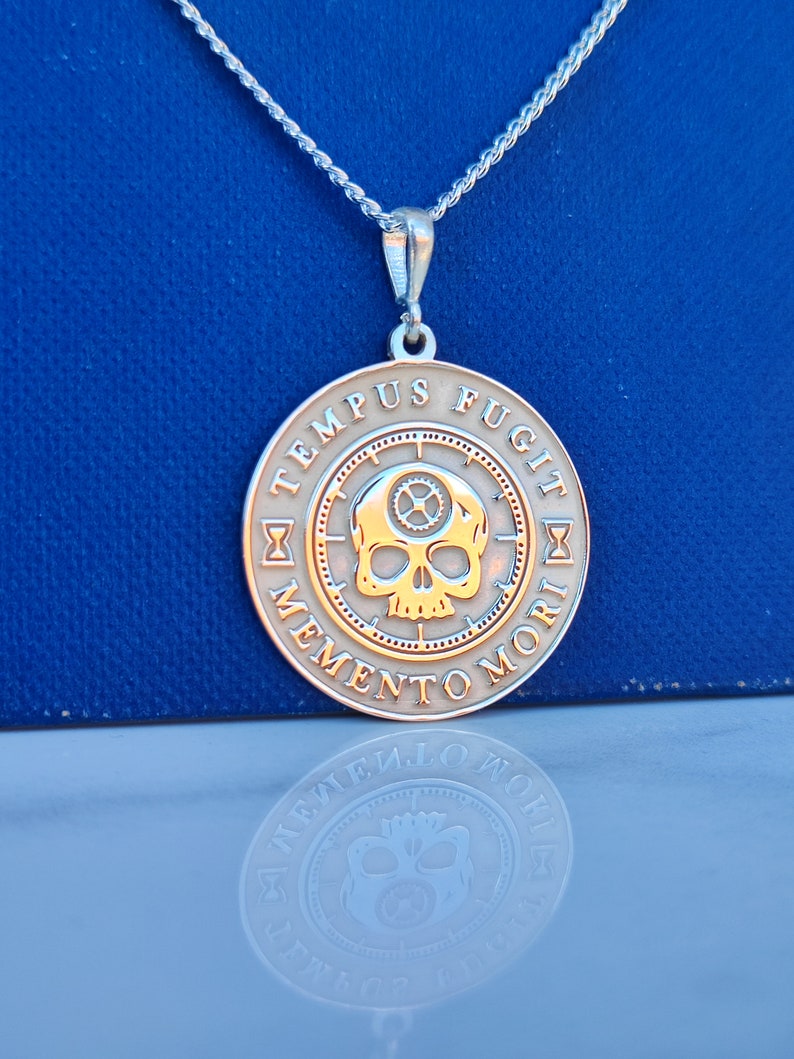 K&ouml;nnte beinhalten: Silberkette mit einem runden Anh&auml;nger, der einen Sch&auml;del mit Zahnr&auml;dern und die Inschrift "TEMPUS FUGIT MEMENTO MORI" in einem Kreis zeigt.