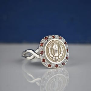 May include: A silver ring with a square-shaped center featuring the Lamar University logo. The logo is a white torch with a laurel wreath and the words "Lamar University" and "Texas". The ring is adorned with red and white gemstones.