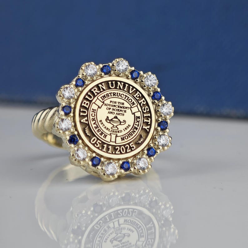 May include: A gold ring with a blue and white stone setting. The ring features the Auburn University seal with the text "Auburn University, Instruction, Research, Extension, For the Advancement of Science and Arts, Established 1856, 05.11.2025".