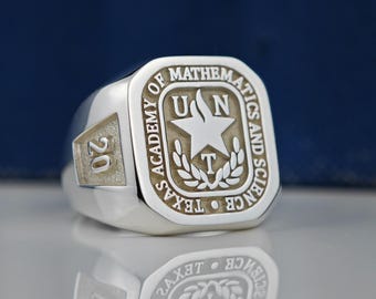 May include: A silver class ring with a square top featuring the inscription "Texas Academy of Mathematics and Science" and the year "20". The ring has a raised star with the letters "UN" above it and a "T" below it.