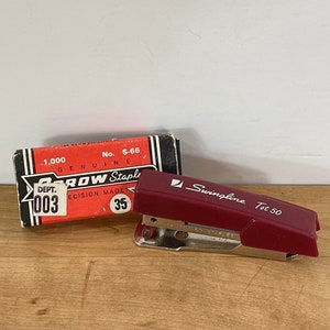 May include: A maroon Swingline Tot 50 stapler and a box of Arrow staples sit on a wooden surface. The stapler has a silver base and the box is red with black and white accents. The box reads "Arrow Staples".