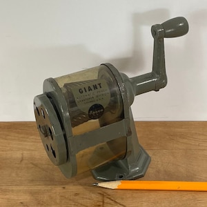 May include: A vintage, gray Giant pencil sharpener with a clear plastic casing and a hand crank. The sharpener is positioned next to a yellow and orange pencil. The text "GIANT" is visible on the sharpener.