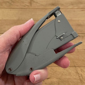 May include: A gray stapler with a silver metal mechanism. The stapler is held in a hand and is set against a wooden background. The stapler is a standard office supply item.