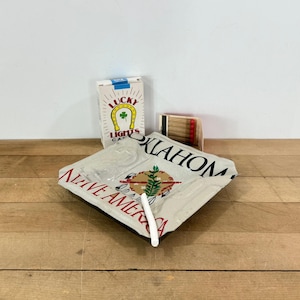 May include: An ashtray with the word "OKLAHOMA" and "NATIVE AMERICA" printed on it, along with a pack of "LUCKY LIGHTS" cigarettes and a box of matches. A white cigarette is resting on the ashtray.