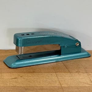 May include: A vintage teal stapler with a silver metal staple holder. The stapler is on a wooden surface. The word "Swingline" is visible on the top of the stapler.