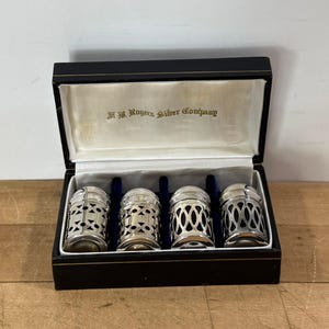 May include: A set of four silver-toned metal shakers with intricate cut-out patterns, housed in a black rectangular presentation box. The interior lid features the gold-printed text F. B. Rogers Silver Company on a white satin lining.