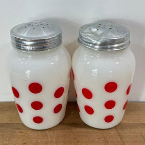 May include: A pair of vintage white glass salt and pepper shakers. Each shaker has a silver metal lid with small holes and is decorated with red polka dots. The shakers are on a wooden surface.