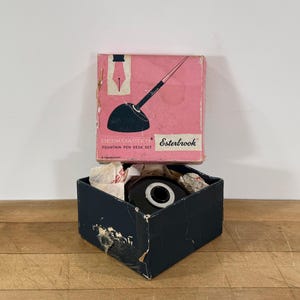 May include: An Esterbrook Deskmaster fountain pen desk set in a pink cardboard box with a black illustration of a pen and base. The set includes a black circular pen base with a silver-toned center, housed inside a dark blue square box.