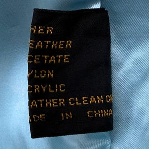 May include: A black clothing tag with gold text, including the words "LEATHER", "ACETATE", "NYLON", and "ACRYLIC". The tag is set against a light blue fabric background, with the words "MADE IN CHINA" visible.