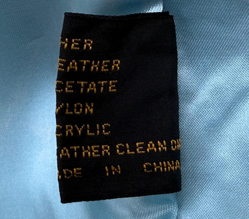 May include: A black clothing tag with gold text, including the words "LEATHER", "ACETATE", "NYLON", and "ACRYLIC". The tag is set against a light blue fabric background, with the words "MADE IN CHINA" visible.