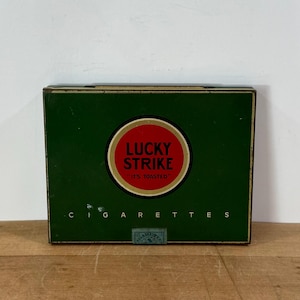 May include: A vintage Lucky Strike cigarette box. The box is green with a red and white circular logo that reads "LUCKY STRIKE" and "IT'S TOASTED". The word "CIGARETTES" is printed in white letters below the logo.