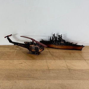 May include: A detailed model of a helicopter and a battleship. The helicopter is black with a red rotor and the battleship is brown. Both models are placed on a wooden surface against a white background.