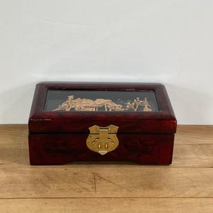 May include: A rectangular, dark red wooden jewelry box with a clear glass top. The top features a gold-colored landscape design. The box has a gold-colored clasp and hinges.