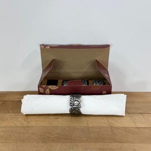May include: A white cloth napkin rolled and secured with a metallic silver-toned napkin ring featuring a detailed embossed fruit and leaf design. Behind the napkin sits an open rectangular cardboard box with a maroon and brown floral pattern.