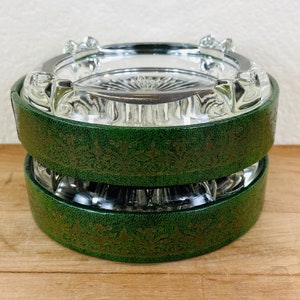 May include: Two clear glass ashtrays stacked, each with a green leather band featuring a gold floral pattern. The ashtrays have a silver rim and are set on a wooden surface.