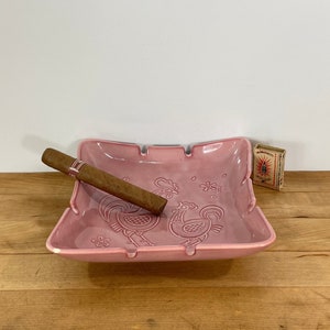 May include: A pink ceramic ashtray with a cigar resting on it. The ashtray features embossed rooster designs and is accompanied by a matchbox. The ashtray is a square shape with curved edges.