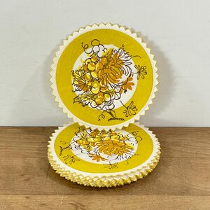 May include: A stack of yellow paper coasters with a scalloped edge. Each coaster features a yellow center with a grape cluster design in shades of yellow and brown. The coasters are stacked on a wooden surface.