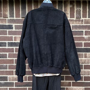 May include: Black suede bomber jacket with a ribbed collar, cuffs, and hem. The jacket has a full-zip closure and two front pockets. The back of the jacket is visible, showing the texture of the suede material. The jacket is paired with a floral shirt and black pants.