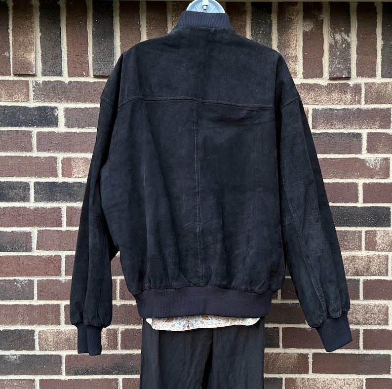 May include: Black suede bomber jacket with a ribbed collar, cuffs, and hem. The jacket has a full-zip closure and two front pockets. The back of the jacket is visible, showing the texture of the suede material. The jacket is paired with a floral shirt and black pants.