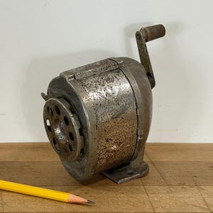 May include: Vintage metal pencil sharpener with a hand crank and a circular blade housing. The sharpener has a worn, rusty appearance, with a wooden handle on the crank. A yellow pencil with a sharpened tip rests on the wooden surface.