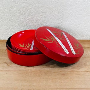 May include: A red lacquered Japanese bento box with a matching lid. The lid and bowls feature gold bamboo and white accents. The box is open, revealing several red bowls inside.