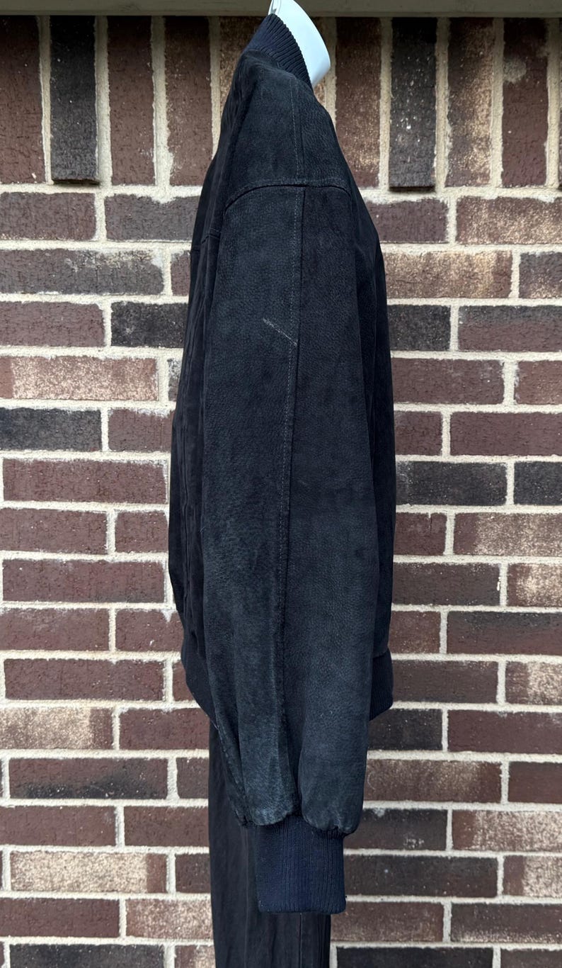 May include: A black bomber jacket with a ribbed collar and cuffs. The jacket is made of a soft, textured material and has a relaxed fit. The jacket is hanging against a brick wall.