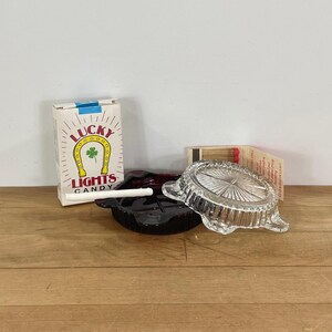 May include: A dark red glass ashtray and a clear glass ashtray with a sunburst pattern, accompanied by a white rectangular box labeled Lucky Lights Candy with a horseshoe graphic and a small open matchbook with red-tipped wooden matches.