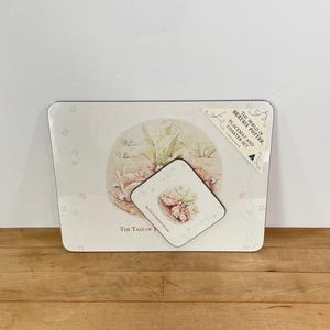 May include: A placemat and coaster set featuring illustrations from Beatrix Potter's stories. The set includes a rectangular placemat and a square coaster, both with a cream background and floral accents. The placemat has a rounded edge.